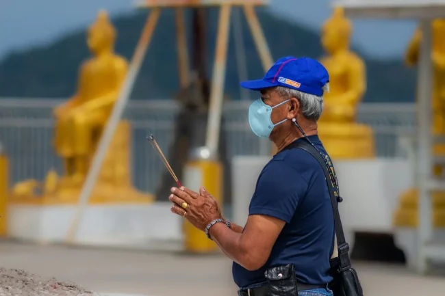 A man wearing a face mask and holding lighted incense sticks. Devotion at Big Buddha, Phuket A man wearing a face mask and holding lighted incense sticks. Devotion at Big Buddha, Phuket